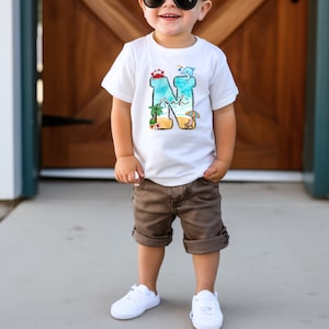 May include: A white t-shirt with a blue, green, and yellow beach scene design featuring a palm tree, beach umbrella, and the letter N. The letter N is filled with the beach scene and the name "Noah" is written inside the letter.