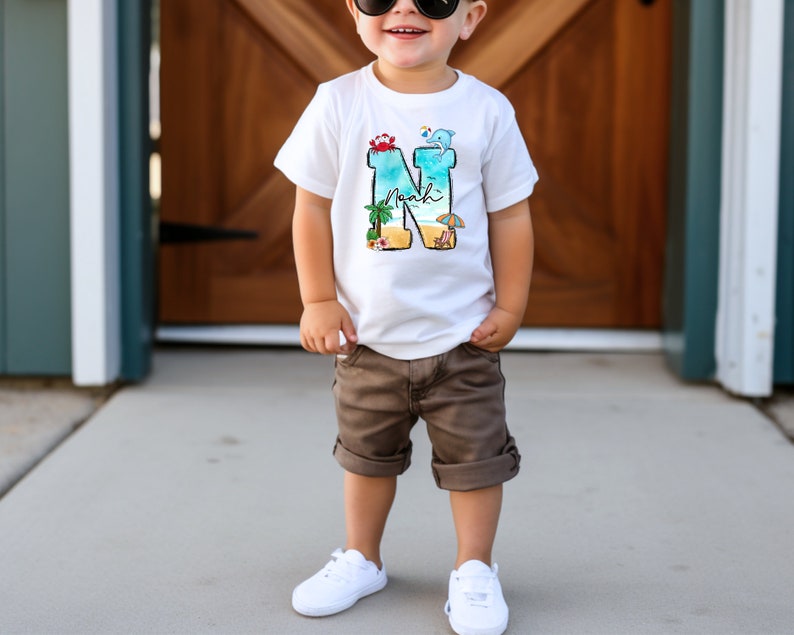 May include: A white t-shirt with a blue, green, and yellow beach scene design featuring a palm tree, beach umbrella, and the letter N. The letter N is filled with the beach scene and the name "Noah" is written inside the letter.