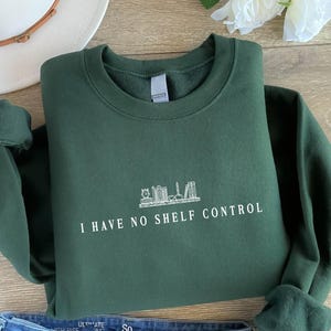May include: Dark green sweatshirt featuring the white text "I HAVE NO SHELF CONTROL" and a skyline graphic. The sleeves are cuffed. A white hat and a flower are in the background. The sweatshirt is folded.