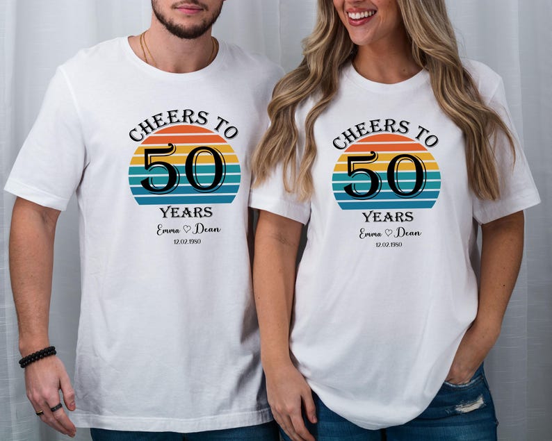 May include: Two white t-shirts with a retro sunset graphic and the text "Cheers to 50 Years". The shirts have the names "Emma" and "Dean" and the date "12.02.1950" printed on them.