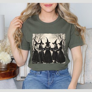 K&ouml;nnte beinhalten: Ein gr&uuml;nes T-Shirt mit einer Vintage-Illustration von f&uuml;nf Frauen, die als Hexen verkleidet sind und ein Banner mit der Aufschrift "Girls Will Be Girls" halten.