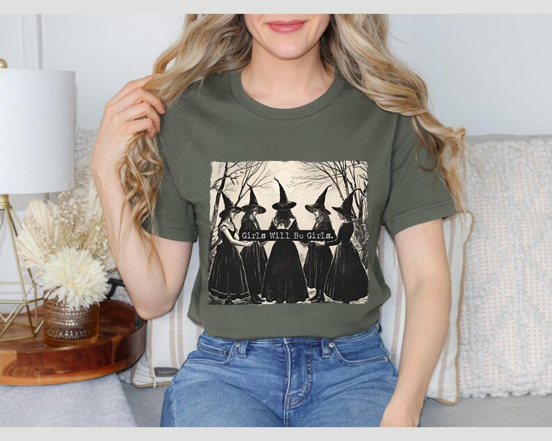 K&ouml;nnte beinhalten: Ein gr&uuml;nes T-Shirt mit einer Vintage-Illustration von f&uuml;nf Frauen, die als Hexen verkleidet sind und ein Banner mit der Aufschrift "Girls Will Be Girls" halten.