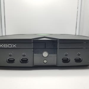 May include: Black Xbox video game console. The console has the word "XBOX" printed on the top left. The console has two controller ports on the front and a power button. The console is sitting on a white surface.
