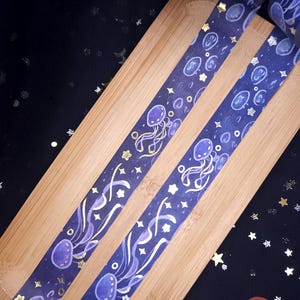 Puede incluir: Dos rollos de cinta adhesiva decorativa. La cinta presenta un fondo azul oscuro con un patrón repetido de medusas moradas, estrellas y burbujas, con detalles dorados. Presentado sobre una superficie de madera.