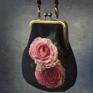 以下が含まれることがあります： キスロック開閉の黒いクラッチバッグ。バラの花柄がプリントされています。バッグはゴールドトーンのフレームと黒いストラップが付いています。バラはピンク色で、背景は暗いです。