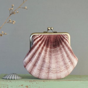 Pode incluir: Uma carteira em forma de concha rosa com fecho de metal. A carteira tem um padrão radial, semelhante a uma concha de vieira. Uma pequena concha está à esquerda da carteira. O fundo é cinza suave.