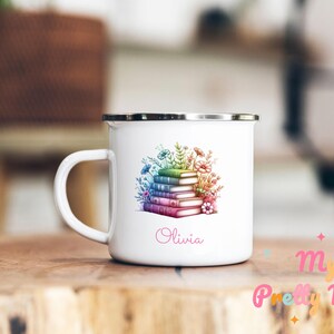 Peut inclure: Mug en émail blanc avec un bord et une anse argentés. La tasse présente une pile de livres aux couleurs de l'arc-en-ciel, entourée de fleurs. Le nom "Olivia" est écrit en rose. La tasse est posée sur une surface en bois.