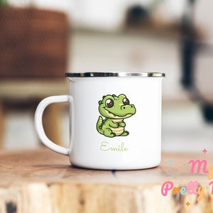 Puede incluir: Taza de esmalte blanco con un cocodrilo verde de dibujos animados con ojos grandes y una sonrisa. La taza tiene un borde y un asa negros. El nombre "Emile" está impreso debajo del cocodrilo.