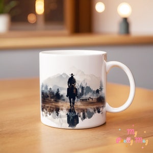 May include: White ceramic mug featuring a colourful image of a cowboy riding a horse through a misty landscape. The image is reflected in a body of water in the foreground.
