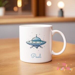 Op de afbeelding: Witte keramische mok met een blauw cartoon UFO-ontwerp en de naam "Paul" in turquoise schrift. De mok heeft een gebogen handvat en staat op een houten oppervlak.