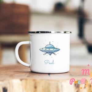 Könnte beinhalten: Weißer Emaillebecher mit silbernem Rand und Henkel. Der Becher zeigt eine blau-graue Cartoon-Illustration eines UFOs mit dem Namen "Paul" in Schreibschrift. Der Becher steht auf einer Holzoberfläche.