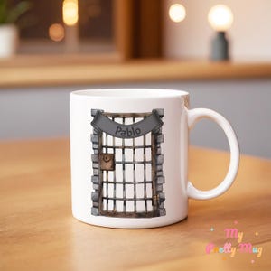 Könnte beinhalten: Weiße Keramik-Tasse mit einer Illustration eines Gefängnistors und dem Namen "Pablo" auf einem Banner. Das Design umfasst ein Schloss, Mauerwerk und einen Holzrahmen. Die Tasse hat einen gebogenen Griff.