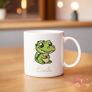 Pode incluir: Caneca de cerâmica branca com uma ilustração de um jacaré verde de desenho animado. O nome "Emile" está escrito em verde abaixo. A caneca tem uma alça curva e está sobre uma superfície de madeira.