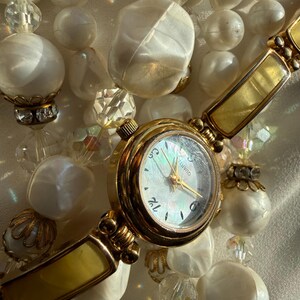 May include: A gold-toned quartz watch with a white face and gold hands, set against a backdrop of white beaded jewelry. The watch has a rectangular band with a yellow-gold finish, and the jewelry features various sizes and shapes of white beads.