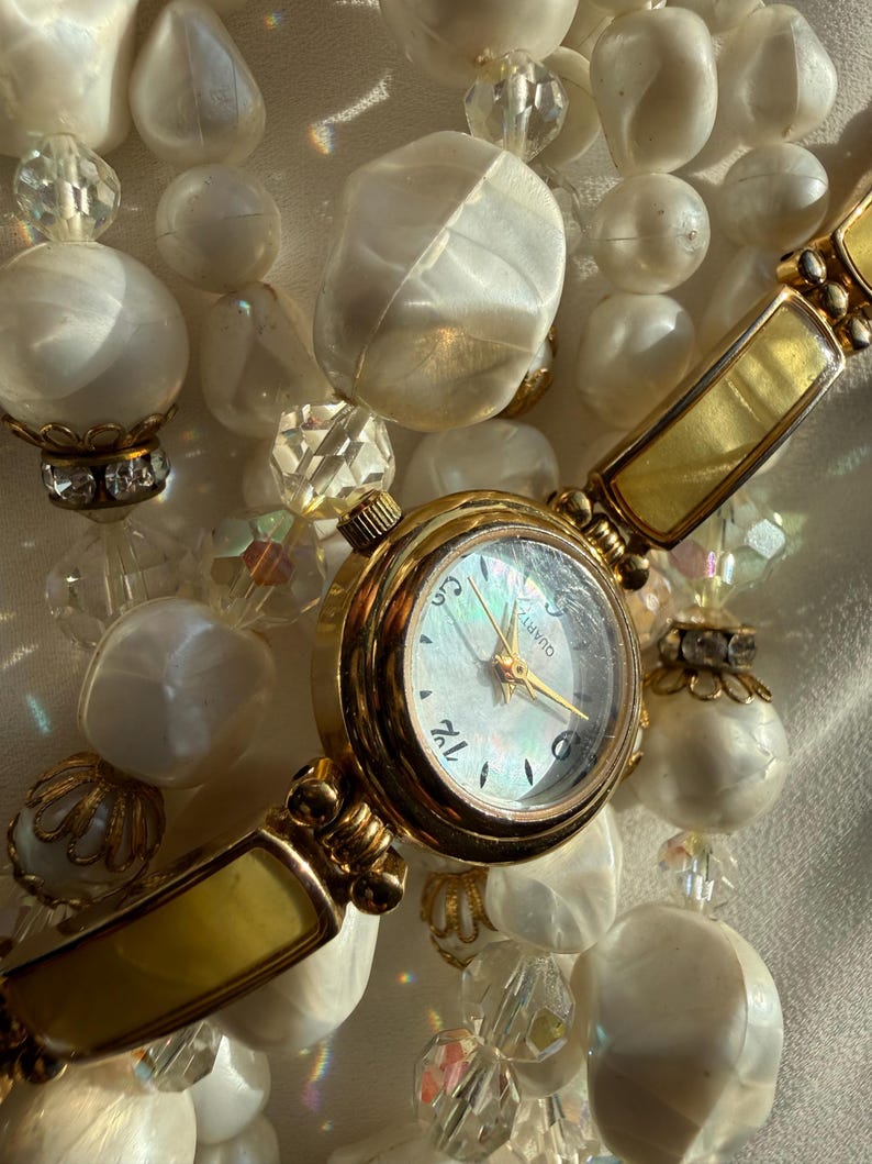 May include: A gold-toned quartz watch with a white face and gold hands, set against a backdrop of white beaded jewelry. The watch has a rectangular band with a yellow-gold finish, and the jewelry features various sizes and shapes of white beads.