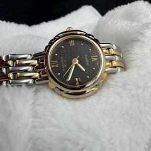 May include: A two-tone wristwatch with a black face and gold hands. The watch has a gold bezel and a silver and gold link band. The watch face features Roman numerals and the text "EXECUTIVE BY JC QUARTZ".