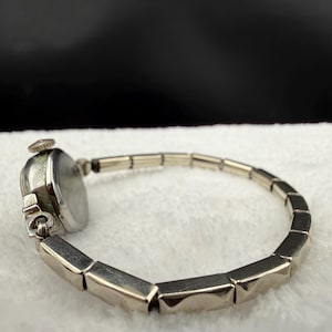 May include: A vintage silver-toned wristwatch with a rectangular face and a linked band. The watch face is round with a light-colored dial. The band is composed of geometric, rectangular links. The watch is set against a white, textured background.