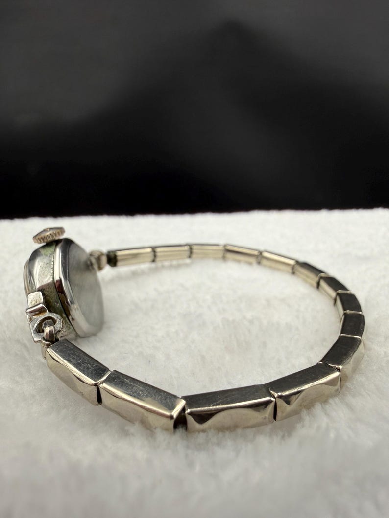 May include: A vintage silver-toned wristwatch with a rectangular face and a linked band. The watch face is round with a light-colored dial. The band is composed of geometric, rectangular links. The watch is set against a white, textured background.