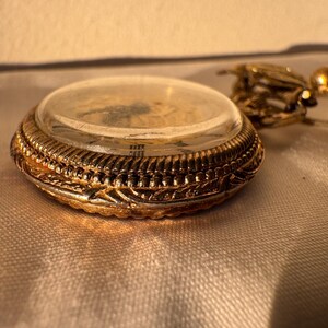 May include: Close-up of a gold-colored pocket watch with a clear glass cover. The watch has intricate detailing around the edges and a delicate chain. The watch is likely an antique or vintage item.