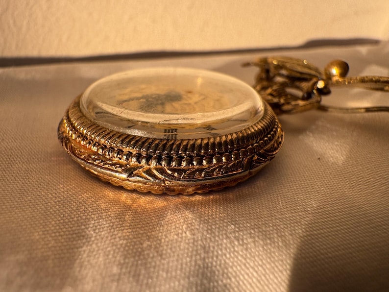 May include: Close-up of a gold-colored pocket watch with a clear glass cover. The watch has intricate detailing around the edges and a delicate chain. The watch is likely an antique or vintage item.