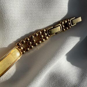 May include: A gold-toned bracelet with a rectangular cream-colored center and a beaded design. The bracelet features a clasp closure and is displayed on a white, textured surface.
