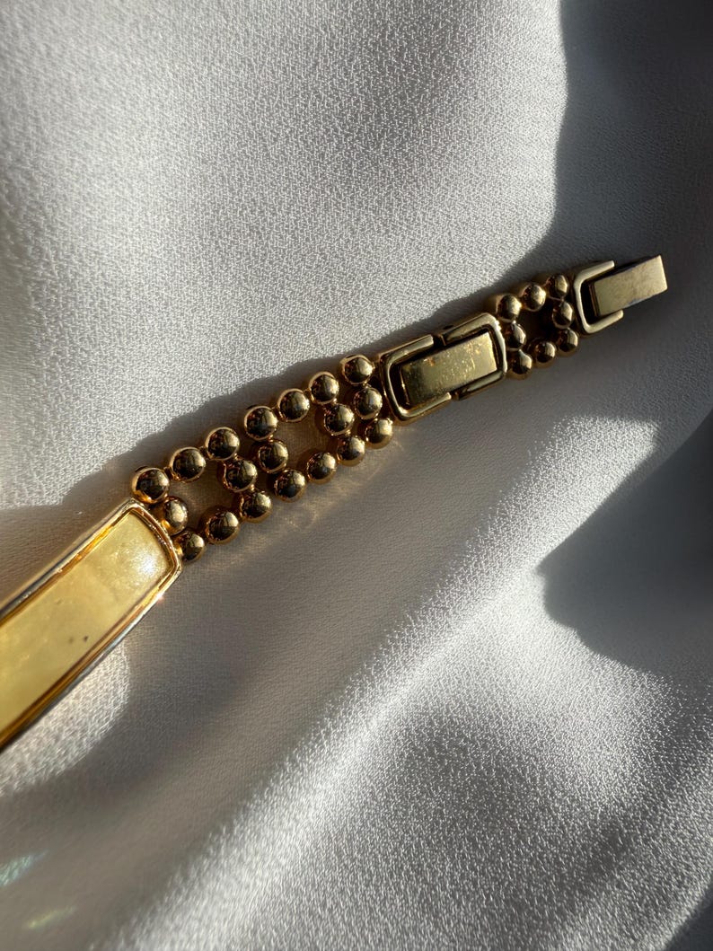 May include: A gold-toned bracelet with a rectangular cream-colored center and a beaded design. The bracelet features a clasp closure and is displayed on a white, textured surface.
