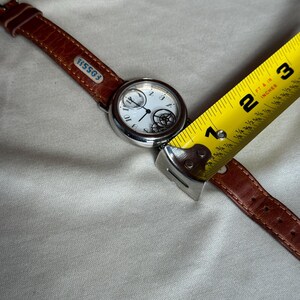 Puede incluir: Un reloj de pulsera Fossil con esfera blanca, caja plateada y correa de cuero marr&oacute;n. La esfera del reloj presenta n&uacute;meros negros y un mecanismo interno decorativo. La correa se mide con una cinta m&eacute;trica amarilla.