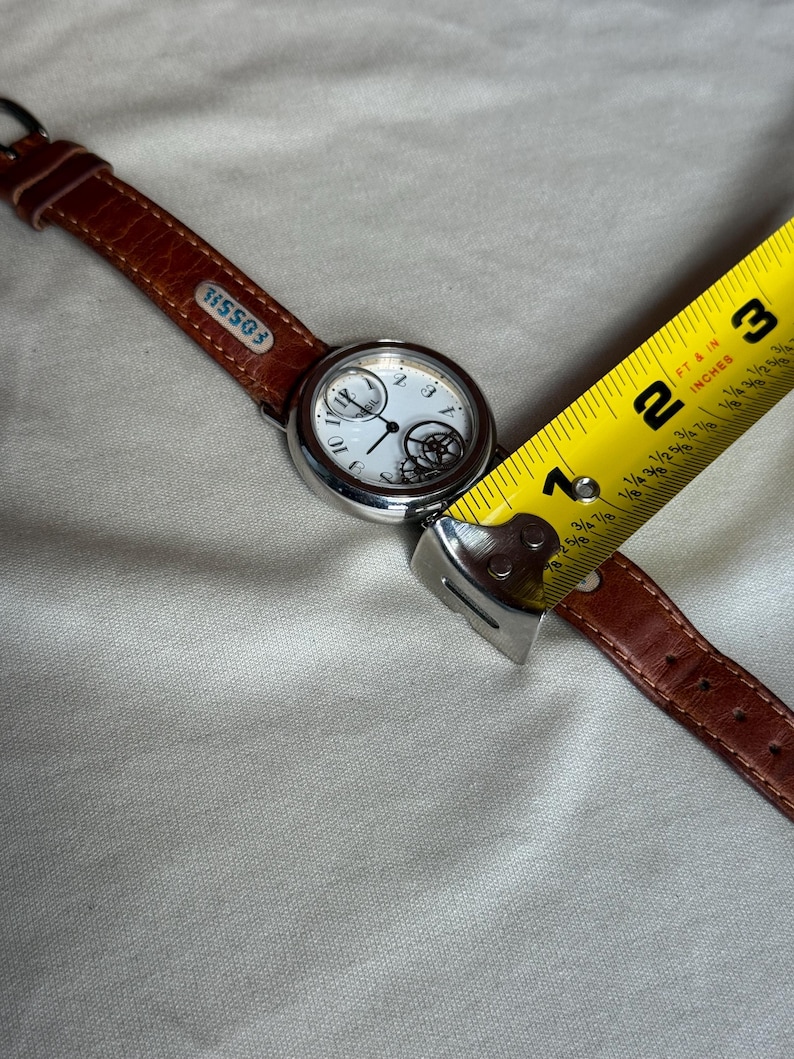 Puede incluir: Un reloj de pulsera Fossil con esfera blanca, caja plateada y correa de cuero marr&oacute;n. La esfera del reloj presenta n&uacute;meros negros y un mecanismo interno decorativo. La correa se mide con una cinta m&eacute;trica amarilla.