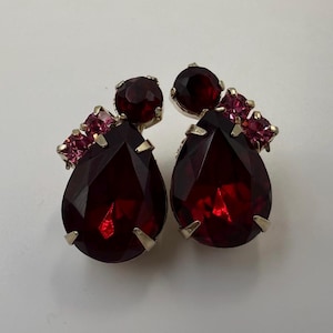 May include: A pair of earrings featuring large, pear-shaped, deep red faceted stones set in silver-toned metal. Each earring is accented with a smaller round red stone at the top and a cluster of three tiny, bright pink square-cut stones.