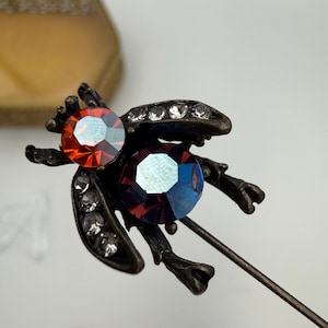 May include: A bronze-toned insect-shaped lapel pin featuring a large round orange faceted crystal for the head and a larger round iridescent blue-purple faceted crystal for the body, accented with small clear crystals along the wings.