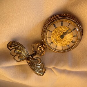 May include: A vintage gold-toned pocket watch with a decorative butterfly-shaped attachment. The watch face features Roman numerals and an ornate gold design. The watch is likely made of metal and is a classic accessory.