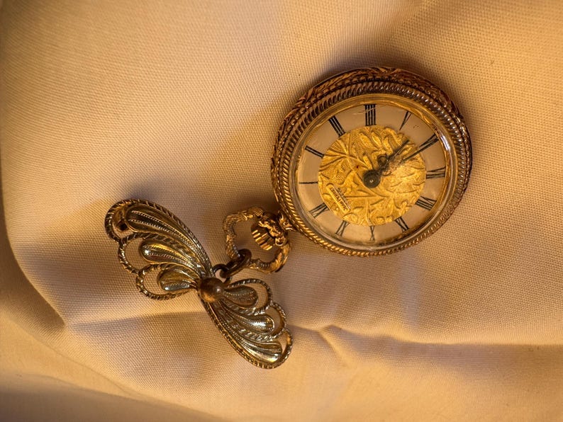 May include: A vintage gold-toned pocket watch with a decorative butterfly-shaped attachment. The watch face features Roman numerals and an ornate gold design. The watch is likely made of metal and is a classic accessory.