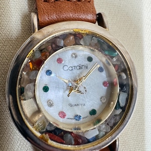 May include: A wristwatch with a brown leather strap and a silver-tone round case. The dial features the text "Carradini" and "QUARTZ JAPAN MOVT" with colorful hour markers. The space between the dial and the crystal is filled with small, loose, multi-colored stones.