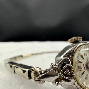 May include: Close-up of a vintage silver-toned wristwatch. The watch features a round face with black numerals and hands. The band is a delicate silver chain with black accents. The watch has a decorative heart-shaped clasp.