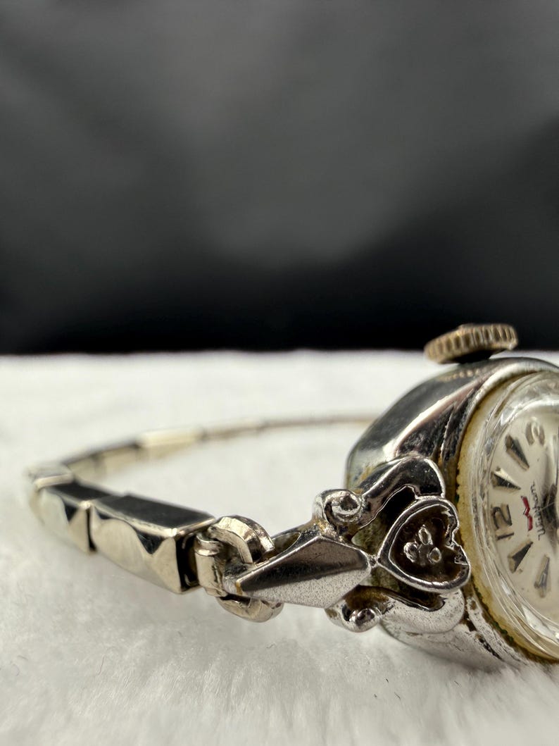 May include: Close-up of a vintage silver-toned wristwatch. The watch features a round face with black numerals and hands. The band is a delicate silver chain with black accents. The watch has a decorative heart-shaped clasp.