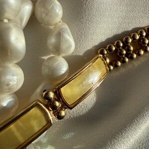 May include: Close-up of a vintage necklace featuring large, off-white, irregular beads and a gold-toned clasp. The clasp has a rectangular design with a light yellow center and small gold ball accents. The necklace is displayed on a cream-colored fabric.