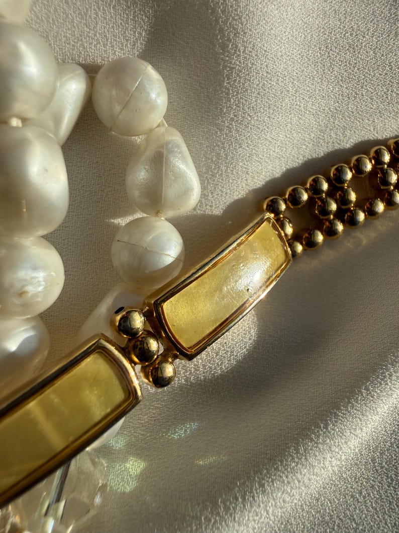 May include: Close-up of a vintage necklace featuring large, off-white, irregular beads and a gold-toned clasp. The clasp has a rectangular design with a light yellow center and small gold ball accents. The necklace is displayed on a cream-colored fabric.