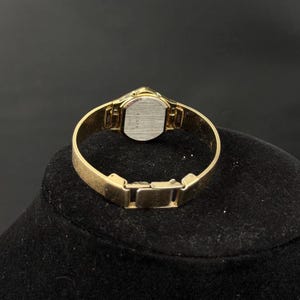 May include: A gold-toned Elgin wristwatch with a rectangular face and a mesh band. The watch has a small, round face with a silver-colored backing. The band is a fine mesh design. The watch is displayed on a black surface.