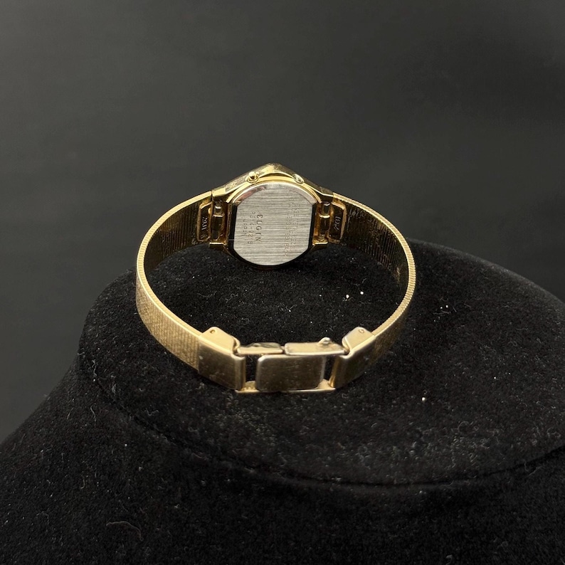 May include: A gold-toned Elgin wristwatch with a rectangular face and a mesh band. The watch has a small, round face with a silver-colored backing. The band is a fine mesh design. The watch is displayed on a black surface.