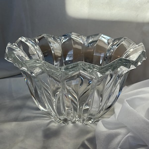 May include: A clear, faceted glass bowl with a scalloped rim. The bowl has a flower-like shape with vertical, leaf-like patterns. The glass reflects light, creating a sparkling effect. The bowl sits on a white surface.