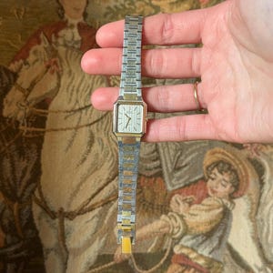 May include: A vintage rectangular wristwatch with a silver and gold-tone metal band and a cream-colored face. The watch has a classic design with a small, square face and a simple, elegant style. The watch band is approximately 6 inches long.