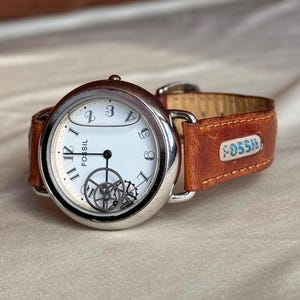 Puede incluir: Un reloj de pulsera Fossil con caja plateada y esfera blanca. El reloj presenta n&uacute;meros y agujas negros, con engranajes visibles. Tiene una correa de cuero marr&oacute;n con el logotipo de Fossil.