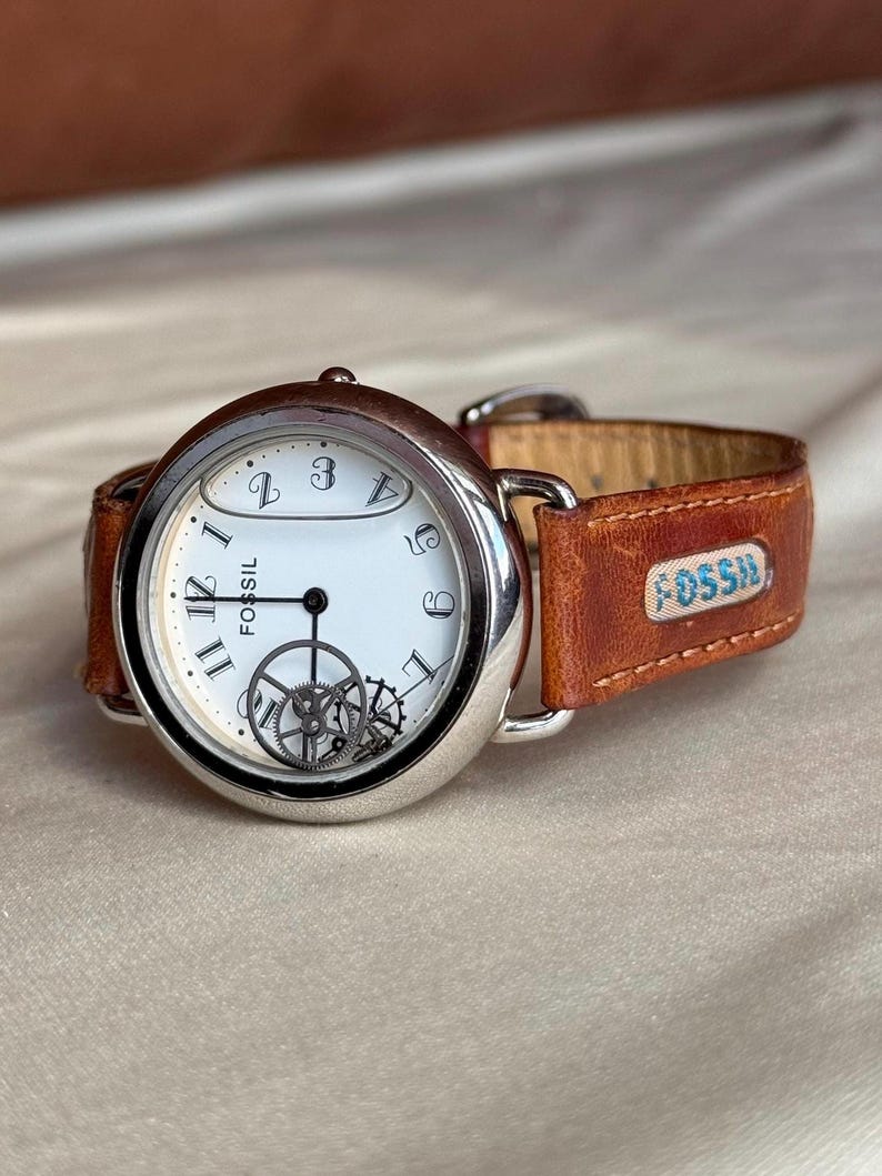 Puede incluir: Un reloj de pulsera Fossil con caja plateada y esfera blanca. El reloj presenta n&uacute;meros y agujas negros, con engranajes visibles. Tiene una correa de cuero marr&oacute;n con el logotipo de Fossil.