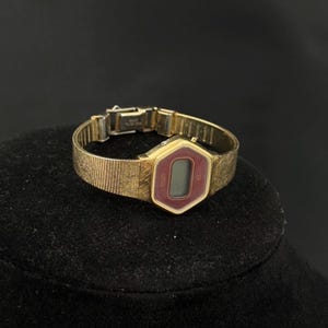 May include: A vintage gold-toned Elgin digital watch with a burgundy display. The watch has a hexagonal face and a textured band. The watch is displayed on a black surface.