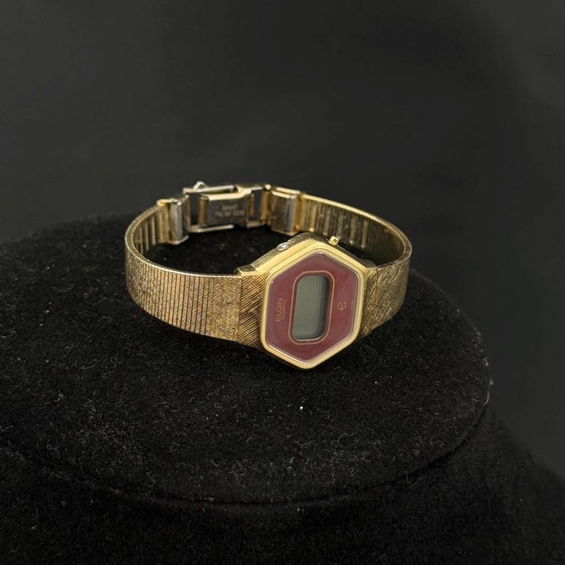 May include: A vintage gold-toned Elgin digital watch with a burgundy display. The watch has a hexagonal face and a textured band. The watch is displayed on a black surface.