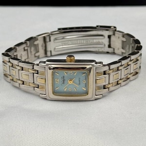 May include: A rectangular wristwatch with a light blue face and gold-tone hands and markers. The watch face has the text "Vanity Fair" and "Quartz". The watch band is silver and gold-tone stainless steel.