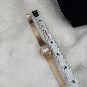 May include: A vintage gold-toned digital watch with a pink face and a mesh band. The watch is next to a Craftsman measuring tape. The watch face displays the time. The watch band is approximately 6 inches long.
