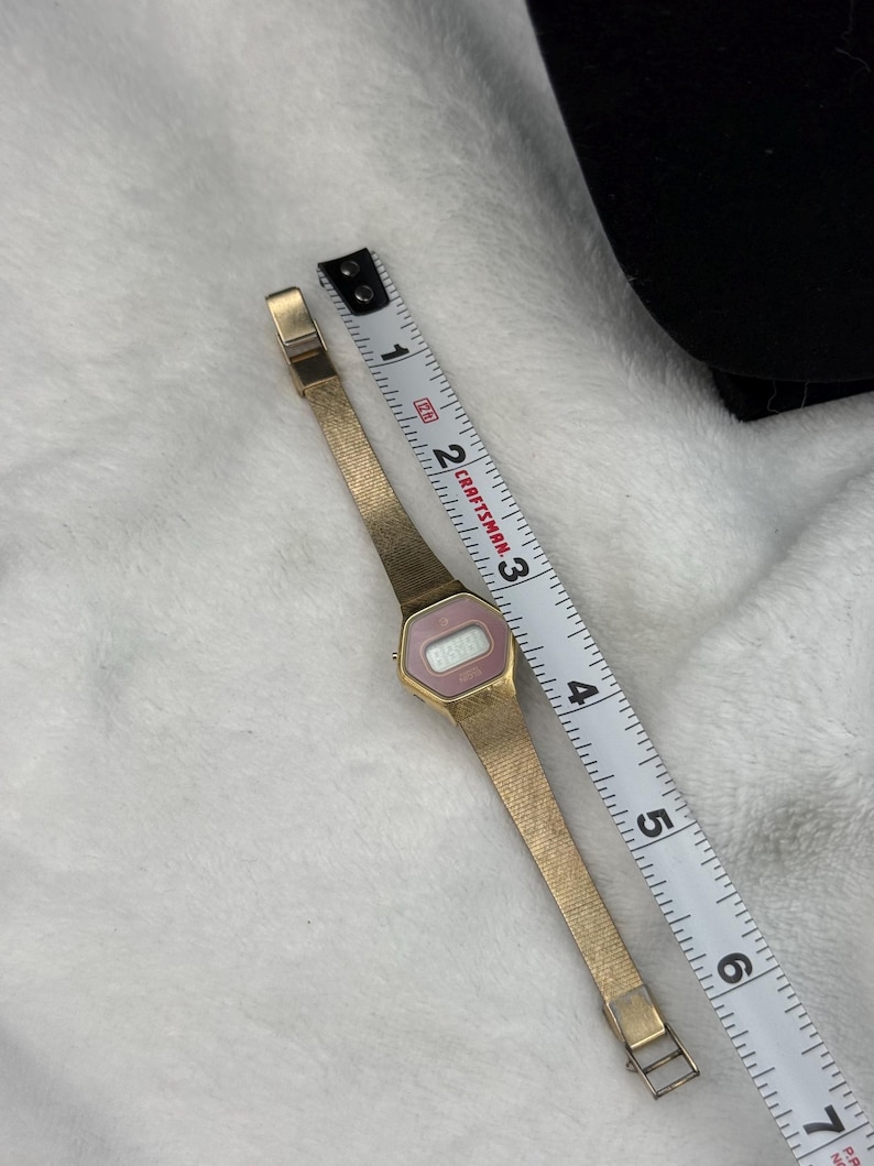May include: A vintage gold-toned digital watch with a pink face and a mesh band. The watch is next to a Craftsman measuring tape. The watch face displays the time. The watch band is approximately 6 inches long.