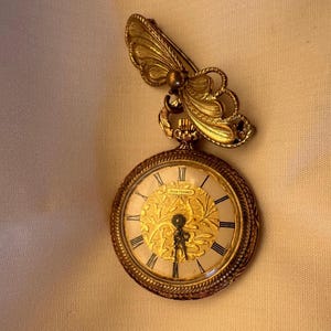 May include: A gold-toned pocket watch with a butterfly brooch. The watch face has Roman numerals and a decorative gold design. The butterfly brooch is attached to the top of the watch.