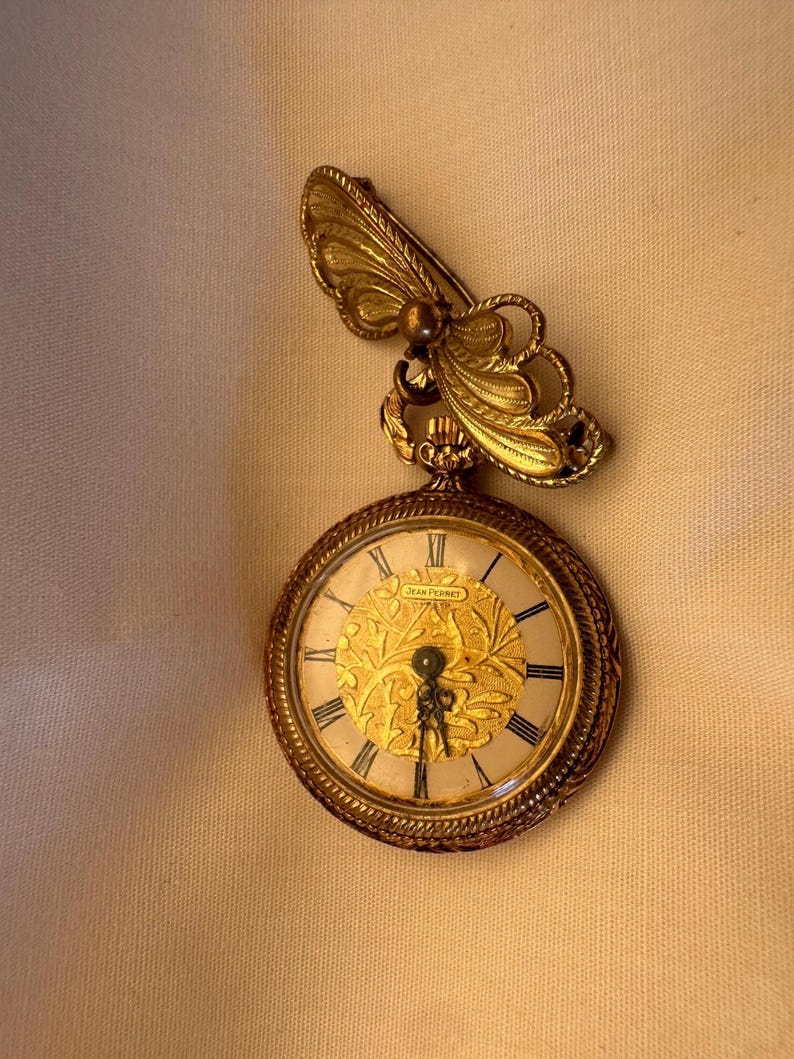 May include: A gold-toned pocket watch with a butterfly brooch. The watch face has Roman numerals and a decorative gold design. The butterfly brooch is attached to the top of the watch.